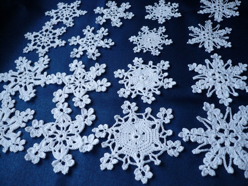 Op de afbeelding: Een verzameling witte gehaakte sneeuwvlokornamenten met ingewikkelde, kantachtige patronen met zesvoudige radiale symmetrie. Elk afzonderlijk stuk toont delicaat draadwerk met gevarieerde geometrische ontwerpen en opengewerkte details.