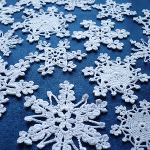 Op de afbeelding: Een verzameling witte gehaakte sneeuwvlokornamenten met ingewikkelde, kantachtige patronen en zespuntige sterontwerpen. Elk afzonderlijk stuk toont delicaat draadwerk dat in een verspreid patroon over een blauw oppervlak is gerangschikt.