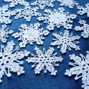 Op de afbeelding: Een verzameling witte gehaakte sneeuwvlokornamenten met ingewikkelde, kantachtige patronen en zespuntige sterontwerpen. Elk stuk is gemaakt met fijn garen en toont delicate lussen en opengewerkte details die in een verspreid patroon liggen.
