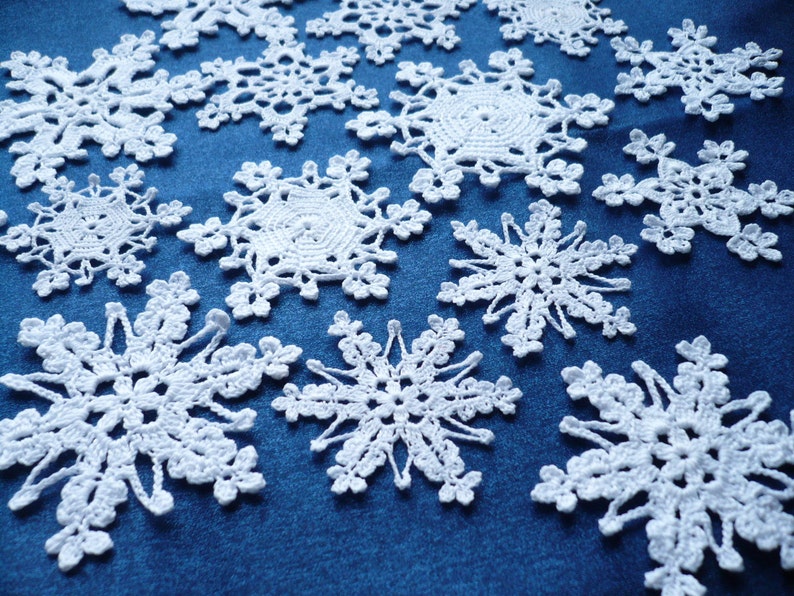 Op de afbeelding: Een verzameling witte gehaakte sneeuwvlokornamenten met ingewikkelde, kantachtige patronen en zespuntige sterontwerpen. Elk stuk is gemaakt met fijn garen en toont delicate lussen en opengewerkte details die in een verspreid patroon liggen.