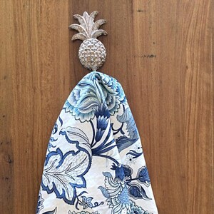 Op de afbeelding: Een decoratieve ananasvormige wandhaak in een metallic afwerking houdt een gevouwen handdoek met een blauw en wit bloemenpatroon vast. De handdoek heeft grote, gestileerde bloemen en bladeren in blauwtinten tegen een witte achtergrond. De haak is op een houten oppervlak gemonteerd.