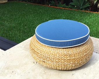 Könnte beinhalten: Ein runder Outdoor-Hocker mit einem Untergestell aus geflochtenen Naturfasern und einem blauen Kissen. Das Kissen ist mit einer weißen Paspel versehen. Der Hocker steht auf einer Steinoberfläche, mit einem grünen Rasen und tropischen Pflanzen im Hintergrund.