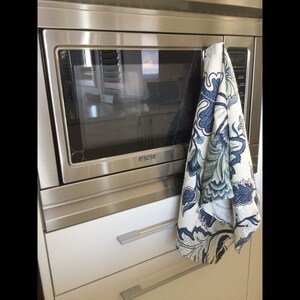 Op de afbeelding: Een roestvrijstalen magnetron met een witte kast eronder. Een decoratieve keukendoek met een blauw en wit bloemmotief hangt aan de magnetronhandgreep.