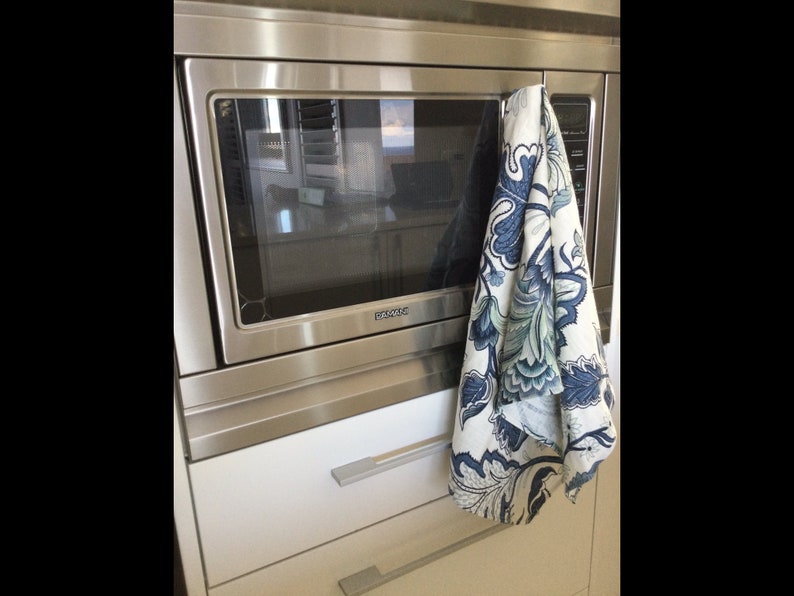 Op de afbeelding: Een roestvrijstalen magnetron met een witte kast eronder. Een decoratieve keukendoek met een blauw en wit bloemmotief hangt aan de magnetronhandgreep.
