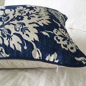 May include: A decorative pillow with a navy blue and cream floral pattern. The pillow has a dark blue trim and a hidden zipper. The pillow is placed on a white bedspread.
