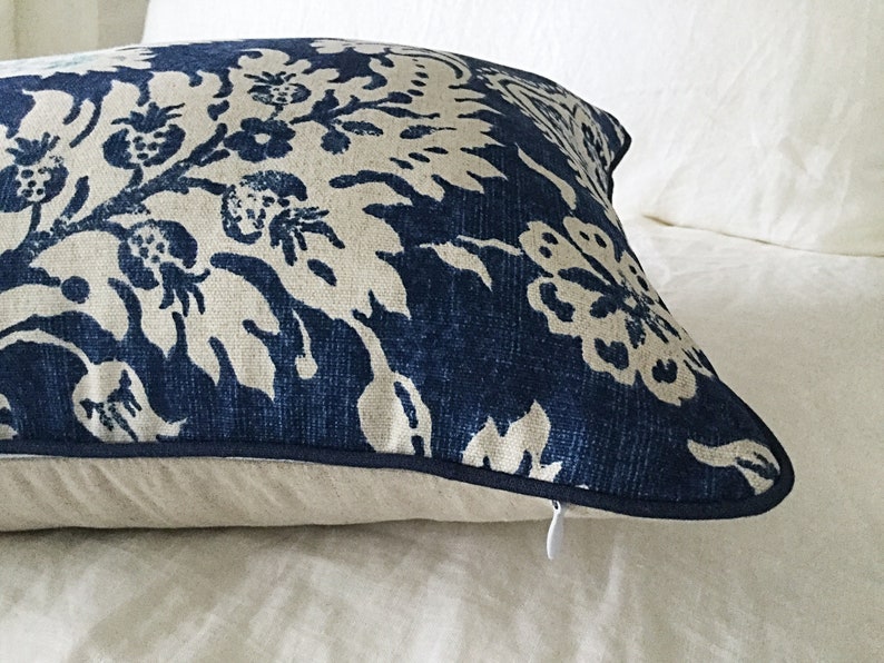 May include: A decorative pillow with a navy blue and cream floral pattern. The pillow has a dark blue trim and a hidden zipper. The pillow is placed on a white bedspread.