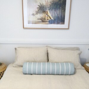May include: A bedroom with a bed featuring a light blue and white striped bolster pillow. The bed has a beige linen duvet and two beige linen pillows. There are two white lamps on nightstands with wicker bases. A framed painting of a sailboat in a tropical setting hangs above the bed.