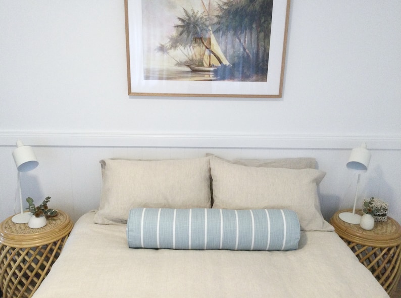 May include: A bedroom with a bed featuring a light blue and white striped bolster pillow. The bed has a beige linen duvet and two beige linen pillows. There are two white lamps on nightstands with wicker bases. A framed painting of a sailboat in a tropical setting hangs above the bed.