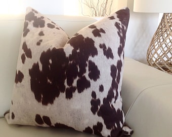 May include: A brown and white cowhide patterned throw pillow. The pillow is on a white couch.