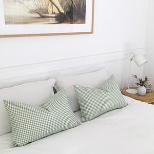 Puede incluir: Una cama blanca con dos almohadas de tiro de cuadros verdes y blancos y una almohada de lino blanca. Una lámpara blanca con una pantalla blanca está sobre una mesita auxiliar de mimbre junto a la cama.