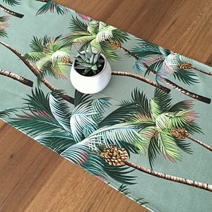 May include: A green table runner with a tropical palm tree print. The runner features a repeating pattern of palm trees with green leaves and brown trunks. A small potted succulent plant sits on top of the runner.