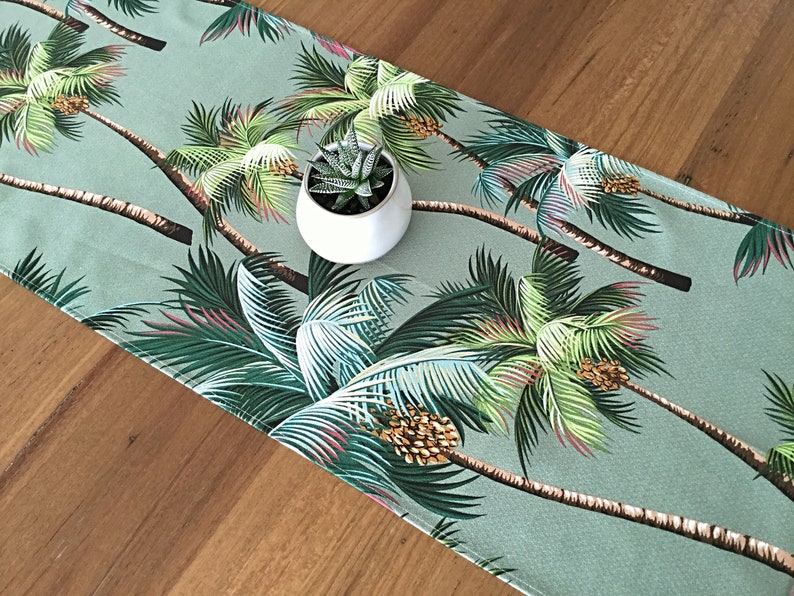May include: A green table runner with a tropical palm tree print. The runner features a repeating pattern of palm trees with green leaves and brown trunks. A small potted succulent plant sits on top of the runner.