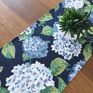 May include: A navy blue table runner with a white and blue hydrangea floral pattern. The runner is on a wooden table with a green plant in a white pot.