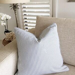 May include: A white and blue striped throw pillow with a subtle sheen. The pillow is on a beige couch with a white lamp in the background.