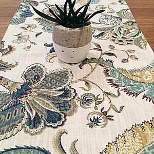 May include: A white table runner with a floral pattern in shades of blue, green, brown, and gold. A small potted succulent plant sits on the runner.