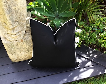 May include: Black pillow with a white piping border. The pillow is sitting on a black wooden surface.