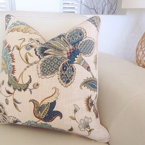 K&ouml;nnte beinhalten: Ein dekoratives Zierkissen mit einem Blumenmuster in Blau-, Gr&uuml;n- und Braunt&ouml;nen auf cremefarbenem Hintergrund. Das Kissen liegt auf einem wei&szlig;en Sofa. Das florale Design zeigt gro&szlig;e, stilisierte Bl&uuml;ten und detaillierte Blattmuster.
