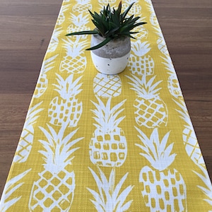 May include: Yellow table runner featuring a white pineapple design. A small succulent plant in a concrete pot is placed on the runner. The runner is made of a textured fabric and is on a wooden table.