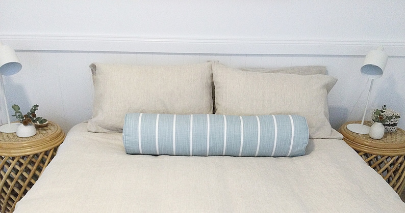 May include: A bed with white linen bedding and two beige pillows. A blue and white striped bolster pillow is placed in the center of the bed.