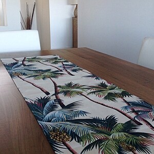 May include: A white table runner with a tropical palm tree print. The runner features green, blue, and brown palm trees on a cream background.