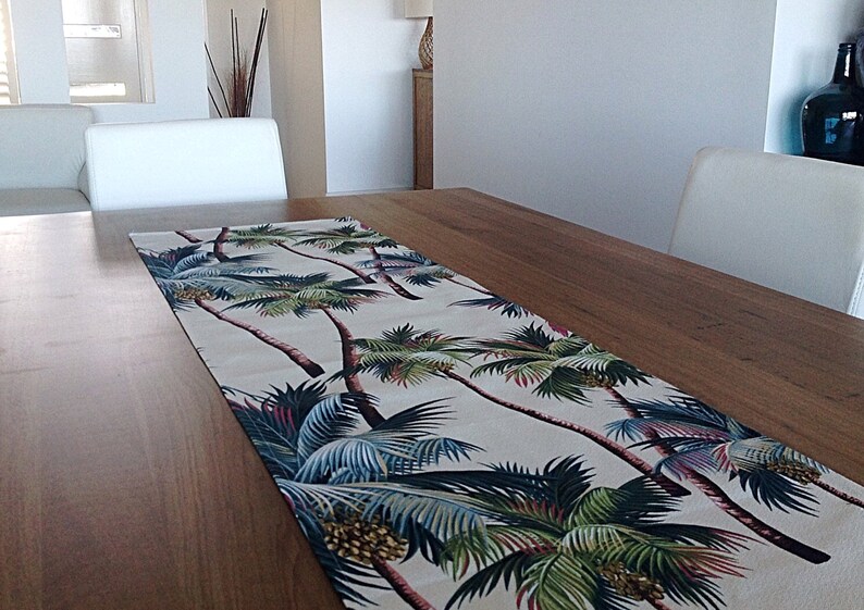 May include: A white table runner with a tropical palm tree print. The runner features green, blue, and brown palm trees on a cream background.
