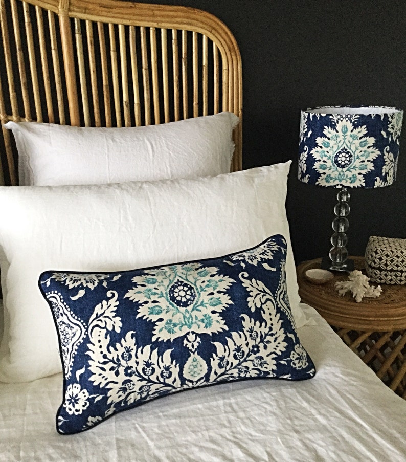 May include: A bedroom scene featuring a decorative pillow with a blue and white floral pattern, a matching lampshade, and white bedding. The headboard is made of woven rattan.