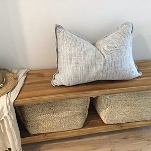 May include: A striped cushion with a dark border sits on a wooden bench. A straw hat with shell details and a fringed cloth are on the bench. Woven baskets are below the bench.