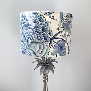 May include: A lampshade with a blue and white floral pattern. The design features large, stylized blooms and foliage in shades of blue against a cream background. The lampshade sits on a silver-coloured lamp base with a pineapple-shaped detail.
