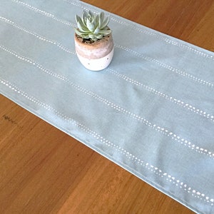 Op de afbeelding: Een lichtblauwe tafelrunner met witte gestippelde lijnen. De runner ligt op een houten tafel met een kleine potplant in het midden.