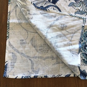 Op de afbeelding: Gevouwen witte stof met een groot blauw bloemenpatroon. Het ontwerp toont gedetailleerde bloemen en bladeren in tinten blauw en blauwgroen. De stof heeft een linnenachtige textuur.