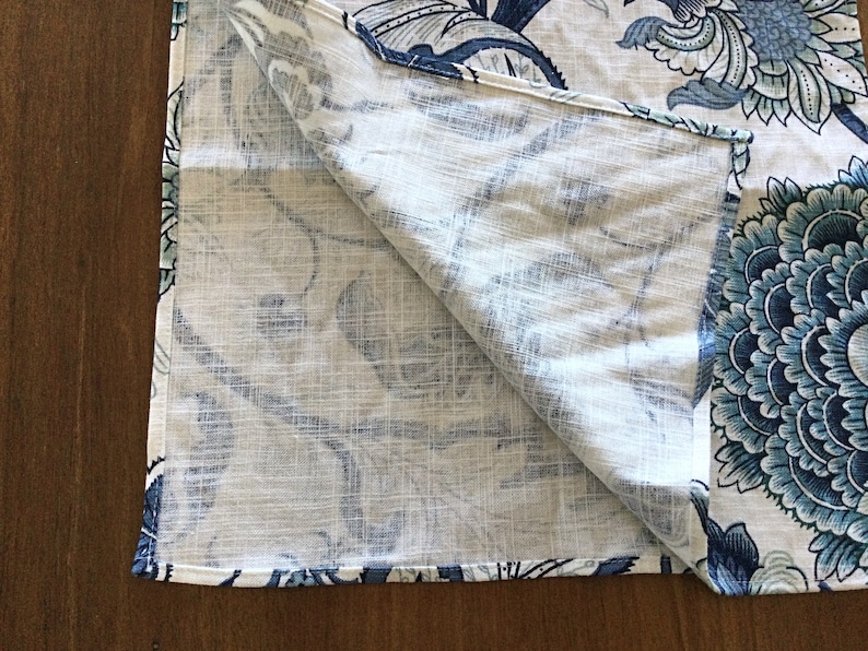 Op de afbeelding: Gevouwen witte stof met een groot blauw bloemenpatroon. Het ontwerp toont gedetailleerde bloemen en bladeren in tinten blauw en blauwgroen. De stof heeft een linnenachtige textuur.