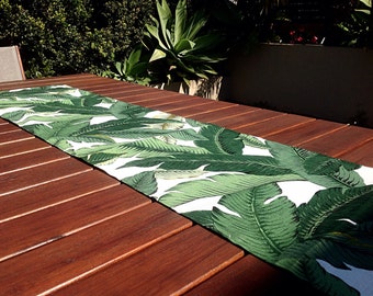 Puede incluir: Un camino de mesa blanco con un patrón de hojas tropicales verdes. El camino de mesa está sobre una mesa de madera.