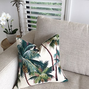 May include: A white throw pillow with a tropical palm tree print in shades of green, brown, and pink. The pillow is on a light gray couch.