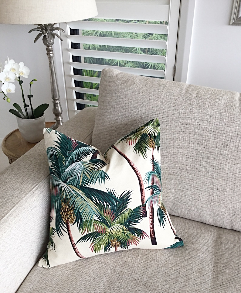 May include: A white throw pillow with a tropical palm tree print in shades of green, brown, and pink. The pillow is on a light gray couch.