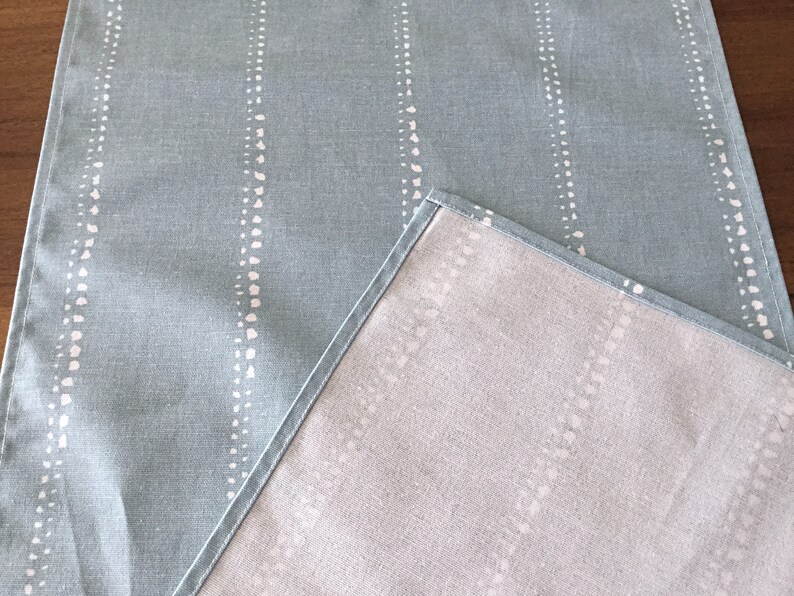 May include: A light blue table runner with a white dotted pattern. The runner is reversible, with a solid light gray color on the other side.