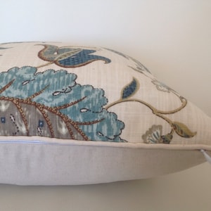 K&ouml;nnte beinhalten: Dekoratives Kissen mit floralem Muster in Blau-, Braun- und Graut&ouml;nen auf cremefarbenem Hintergrund. Das Kissen hat eine einfarbige cremefarbene Basis und einen Rei&szlig;verschluss. Das Design zeigt stilisierte Blumen und Bl&auml;tter.