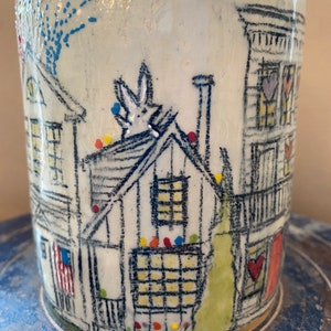 May include: White ceramic mug with a hand-painted design of a small town with houses, a rabbit on a roof, and a flag. The houses have windows and doors, and the roof of one house has a chimney. The mug has a speckled glaze and a light blue rim.
