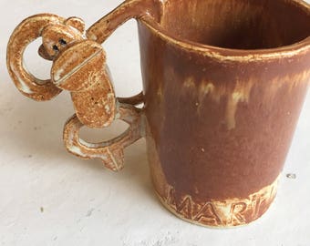 May include: A brown ceramic mug with a monkey-shaped handle. The mug has a light brown glaze and the word "MAR" is visible on the bottom.