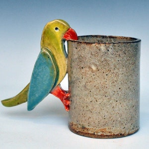 May include: A ceramic mug with a parrot-shaped handle. The parrot is yellow, green, and blue with red accents. The mug is a speckled brown color.