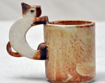 Könnte beinhalten: Eine Keramiktasse mit einem katzenförmigen Henkel. Die Tasse ist in einem hellbraunen Ton glasiert mit dunkleren braunen Akzenten. Der Katzenhenkel ist weiß mit braunen Akzenten.