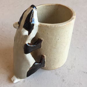 May include: A ceramic mug with a badger figurine attached to the side. The mug is beige with a speckled glaze. The badger is white with black markings and is holding onto the mug with its paws.