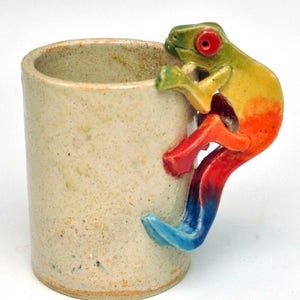 May include: A ceramic mug with a colorful frog handle. The mug is beige with a speckled glaze. The frog is green, yellow, orange, red, and blue.