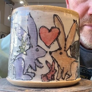 May include: A ceramic mug with a cream-colored base and rim, featuring a hand-drawn design of rabbits and a heart. The rabbits are in shades of purple, orange, and red, with black outlines. The mug is cylindrical and appears handmade.