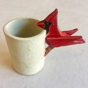 May include: A white ceramic mug with a red cardinal bird handle. The cardinal is perched on the side of the mug and has its wings spread out.