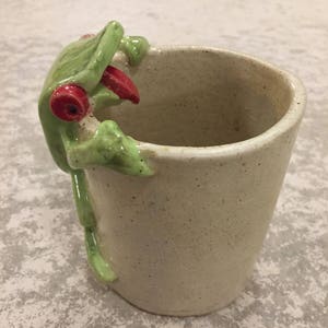 May include: A ceramic mug with a green frog figurine attached to the side. The frog has red eyes and a long tongue sticking out. The mug is white with a speckled glaze.