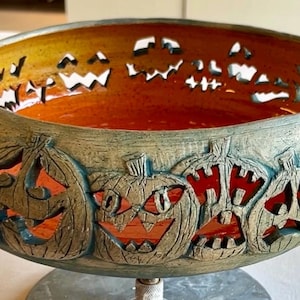 May include: A round ceramic bowl featuring a textured, weathered green exterior with carved jack-o'-lantern faces. The interior of the bowl is a smooth, vibrant orange, and the cut-out pumpkin designs allow light to pass through the walls.