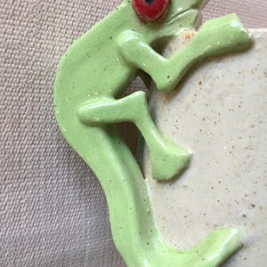 May include: A green ceramic frog figurine with a red eye, climbing up a white ceramic vase.