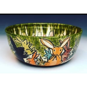 May include: A green ceramic bowl with a whimsical design of rabbits and other woodland creatures. The bowl is hand-painted with details in black, orange, yellow, and blue.