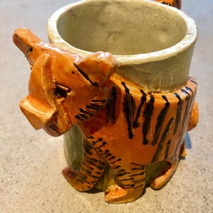 May include: A ceramic mug shaped like a tiger. The mug is orange and black with a white interior. The tiger's head is the handle of the mug.