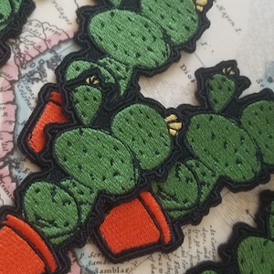May include: A set of three embroidered cactus patches featuring green cacti and orange pots. The patches are displayed on a vintage map background.